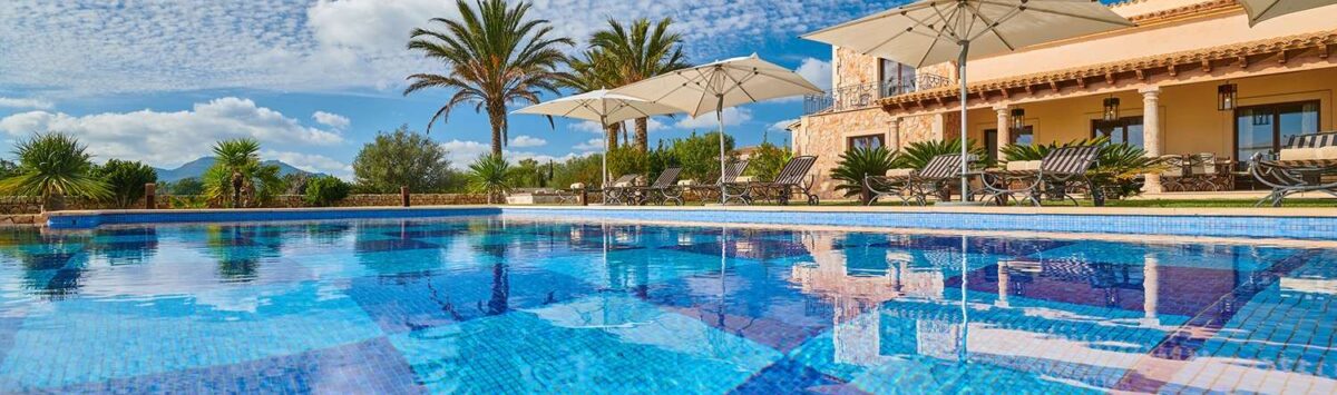 Large outdoor pool with mosaic tiles, sun loungers and parasols in front of a Mediterranean finca in Portocolom
