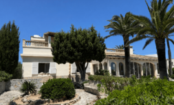 Außenansicht einer mediterranen Villa in Manacor mit Palmen, Arkaden, Balustrade und gepflegtem Garten bei blauem Himmel.
