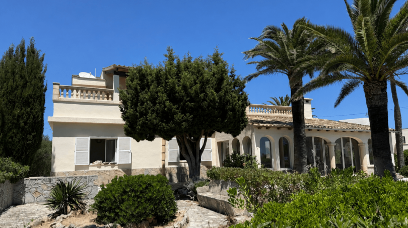 Exterior view of a Mediterranean villa in Manacor with palms, arcades, balustrade and well-maintained garden under blue sky.