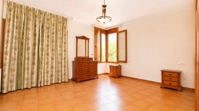 Helles Schlafzimmer der Finca in Marratxí mit Terrakottaboden, Holzmöbeln und floralem Vorhang.