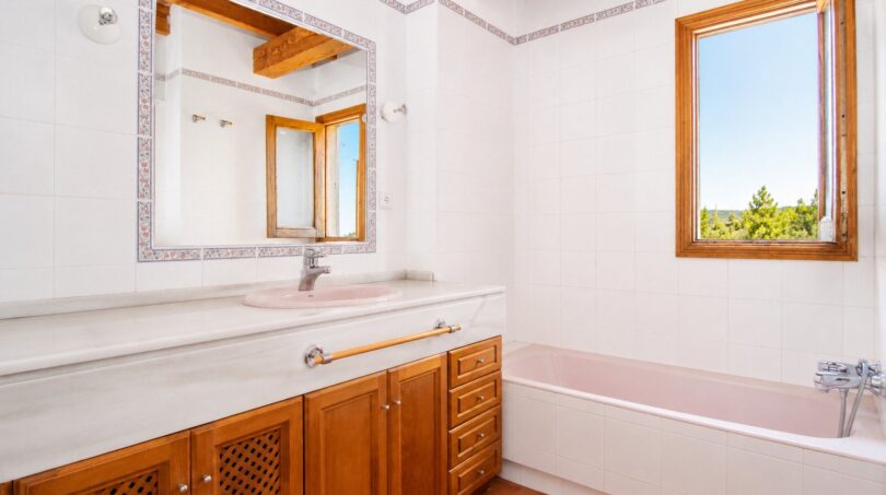 Helles Badezimmer der Finca in Marratxí mit Holzunterschrank, rosa Badewanne und Fenster mit Naturblick.
