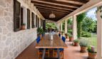 Terraza cubierta de la finca en Lloseta con mesa larga de madera, sillas azules, pared de piedra y vista al jardín.
