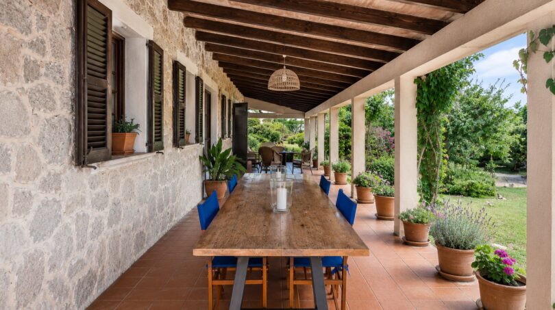 Terraza cubierta de la finca en Lloseta con mesa larga de madera, sillas azules, pared de piedra y vista al jardín.