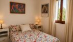 Dormitorio luminoso de la finca Lloseta con cama doble, colcha con flores, grabados artísticos y ventanas de madera con vista al jardín.