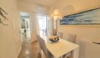 Bright dining room with white table, gray chairs, flower lamp and seascape painting in Portocolom apartment