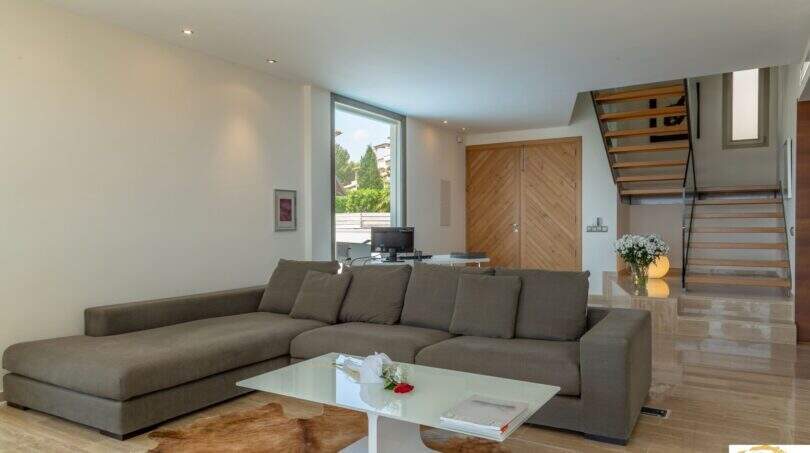 Sala de estar luminosa de la villa de lujo en Porto Cristo con sofá en L, escalera de cristal y puerta de madera