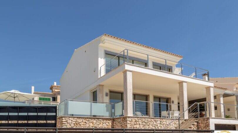 Villa de lujo mediterránea Cala Magrana: Fachada exterior blanca con balcón de cristal, muro de piedra natural y terraza en primera línea de mar.