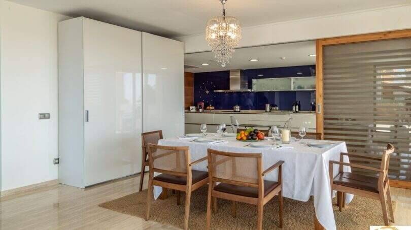 Elegante comedor con lámpara de cristal, mesa dispuesta y cocina de diseño abierta de la villa de lujo en Cala Magrana.