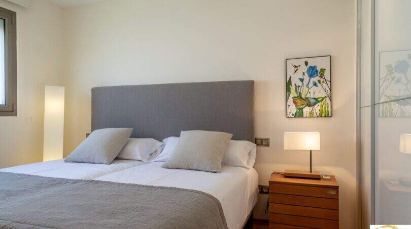 Elegante dormitorio de la villa de lujo en Cala Magrana con cama doble, cabecero tapizado gris y mesilla de madera