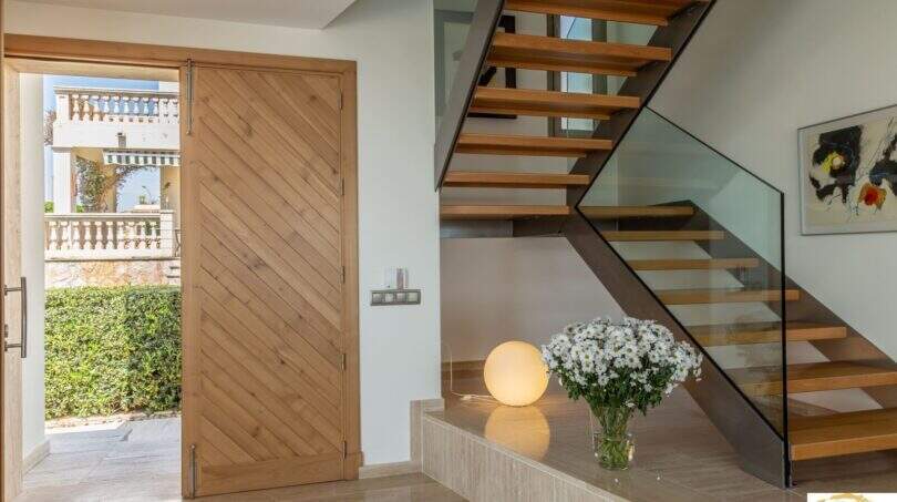 Zona de entrada de la villa de lujo en Cala Magrana con escalera de madera, barandilla de cristal y elegante suelo de piedra natural.