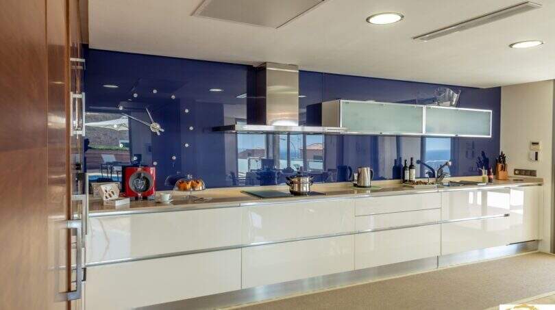 Cocina de lujo moderna con frentes de alto brillo blancos, salpicadero de cristal azul y vistas al mar en Villa Cala Magrana