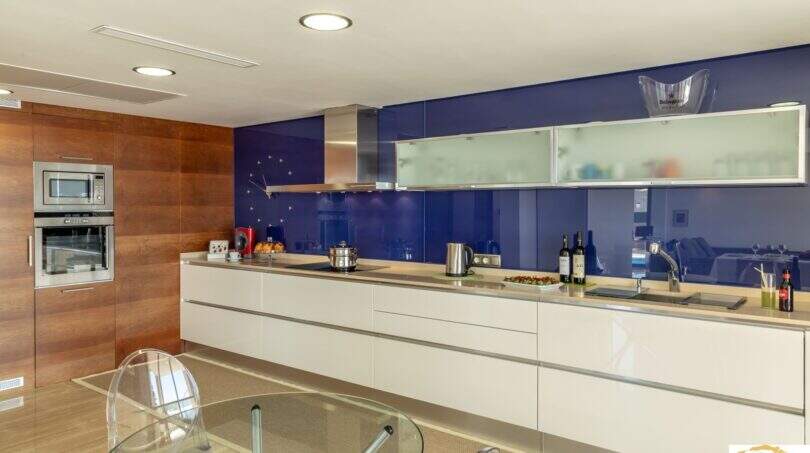 Cocina de lujo moderna con salpicadero de cristal azul, frentes de alto brillo blancos y elementos de madera en Villa Cala Magrana