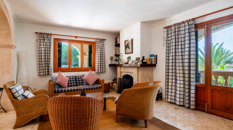 Salón de la villa de Cala Murada con chimenea, muebles de ratán, cortinas a cuadros y acceso a la terraza