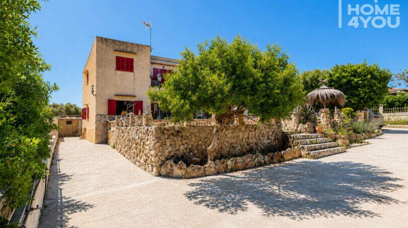 Außenansicht Finca in Cala Llombards mit Natursteinmauer, roter Fensterläden und mediterranem Baumbestand
