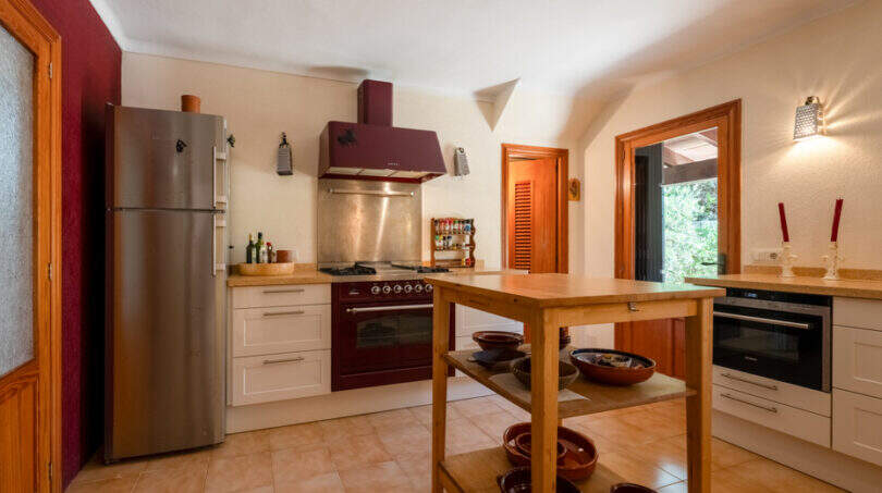 Amplia cocina de la villa de Cala Murada con isla de cocina, cocina de color burdeos, frigorífico de acero inoxidable y cuencos rústicos de terracota.