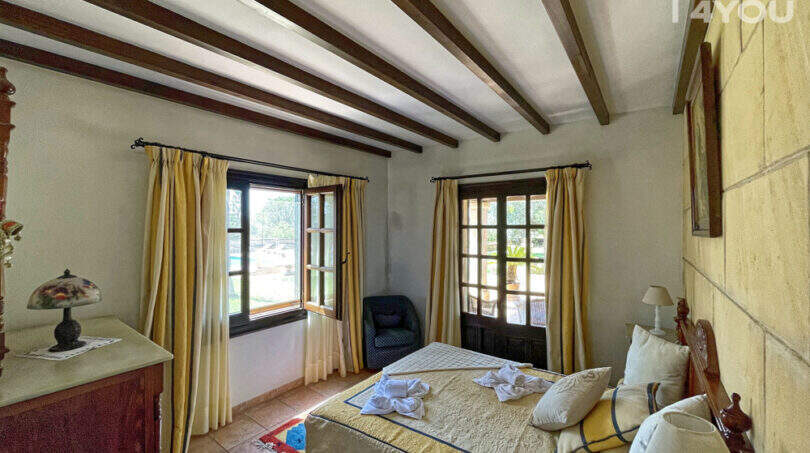 Schlafzimmer der Finca mit Holzbalkendecke, Doppelbett, gelben Vorhängen und Blick auf Pool und Garten.