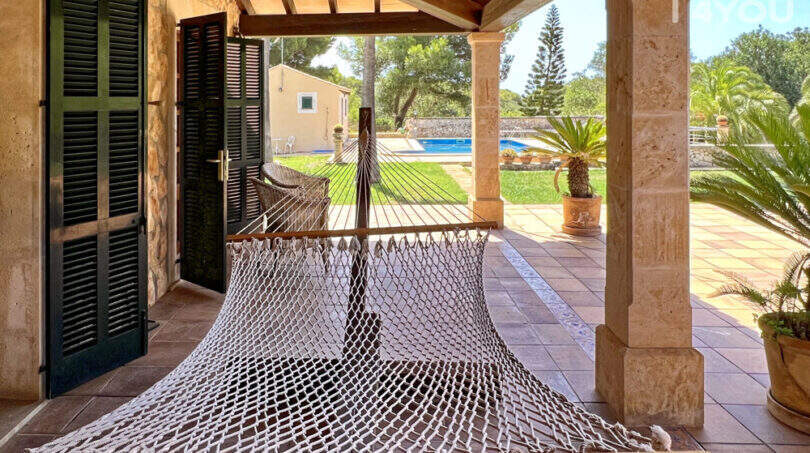 Überdachte Terrasse der Finca mit Hängematte, Natursteinboden und Blick auf Pool und Garten.