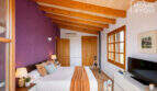 Schlafzimmer der Finca Ses Salines mit Holzbalkendecke, lila Akzentwand und mediterranem Charme.