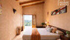 Schlafzimmer der Finca in Ses Salines mit Holzbalkendecke, Doppelbett und Balkonblick ins Grüne.