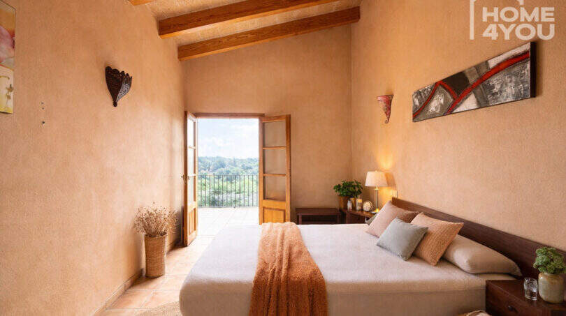 Schlafzimmer der Finca in Ses Salines mit Holzbalkendecke, Doppelbett und Balkonblick ins Grüne.