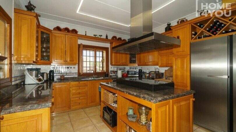 Mediterrane Finca: Geräumige Küche mit Holzschränken, Kochinsel, Granitarbeitsplatten und Weinregal.