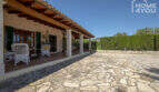 Überdachte Terrasse der Finca Lloseta mit Natursteinboden, Holzbalken & weißen Rattanmöbeln, Gartenblick