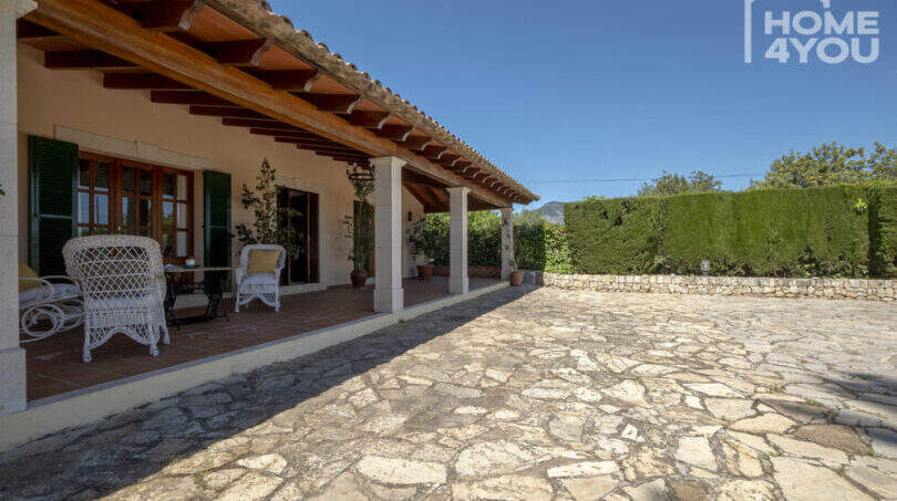 Überdachte Terrasse der Finca Lloseta mit Natursteinboden, Holzbalken & weißen Rattanmöbeln, Gartenblick