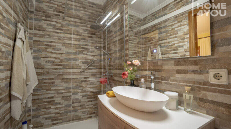 Modernes Bad der Finca Lloseta mit Natursteinoptik-Fliesen, Glasdusche, Aufsatzwaschbecken und Blumendeko.