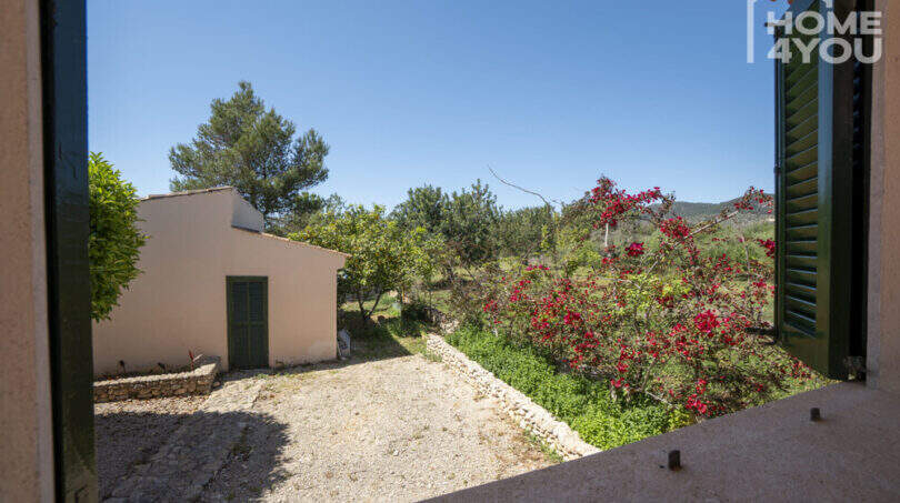 Blick aus Fenster auf mediterranen Innenhof mit Nebengebäude, roter Bougainvillea und grüner Landschaft in Lloseta