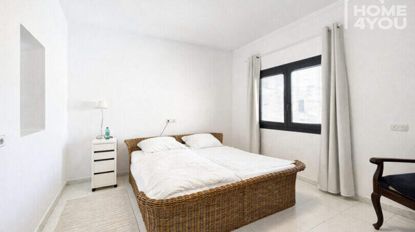 Helles Schlafzimmer mit Rattanbett, weißer Bettwäsche, Nachttisch und schwarzem Fenster im Ibiza-Stil
