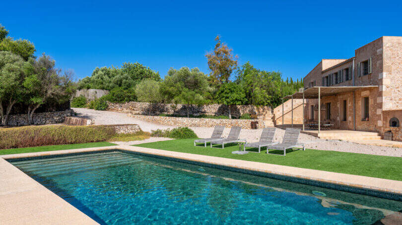 Finca Esporles: Großer Pool mit Liegestühlen, Natursteinhaus und mediterranem Garten unter blauem Himmel.