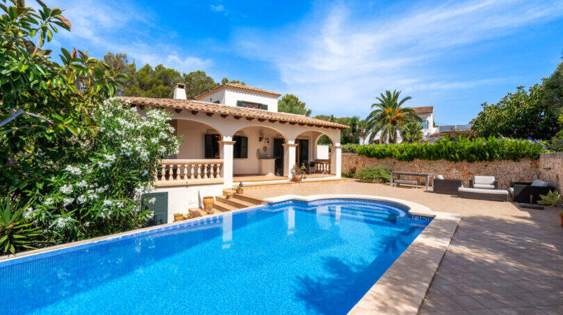 Villa mediterránea en Cala Murada con una gran piscina rectangular, terraza y una exuberante vegetación bajo un cielo azul.