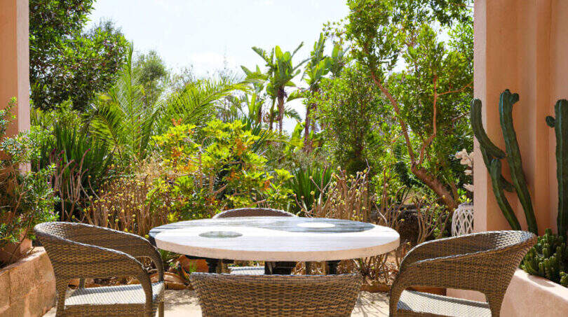 Überdachte Terrasse mit Rattanmöbeln und Marmorrundetisch, umgeben von üppiger mediterraner Gartenvegetation beim Agroturismo Ses Salines