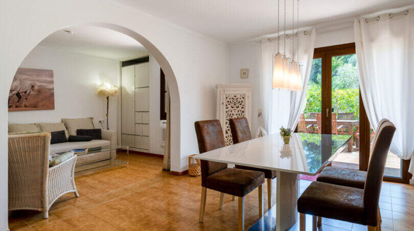 Helles Esszimmer mit weißem Tisch, braunen Stühlen, Rundbogen zum Wohnzimmer und Terrassenzugang in Cala Murada