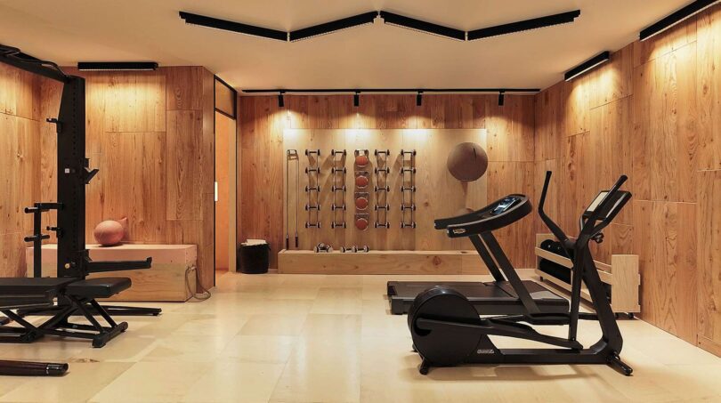 Moderner Fitnessraum der Luxusfinca mit Holzverkleidung, Laufband, Crosstrainer und Wandhalterungen für Medizinbälle.