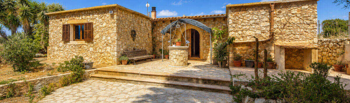 Rustikale Naturstein-Finca in Felanitx mit gepflastertem Innenhof, Brunnen und mediterranem Bewuchs unter blauem Himmel.