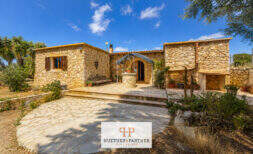 Rustic finca in Felanitx: natural stone building with courtyard, well and Mediterranean planting under blue sky.