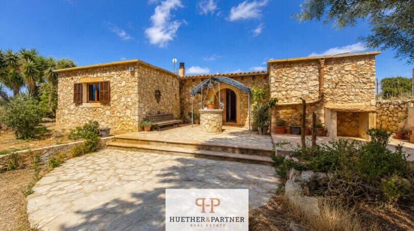 Rustikale Naturstein-Finca in Felanitx mit gepflastertem Innenhof, Brunnen und mediterranem Bewuchs unter blauem Himmel.