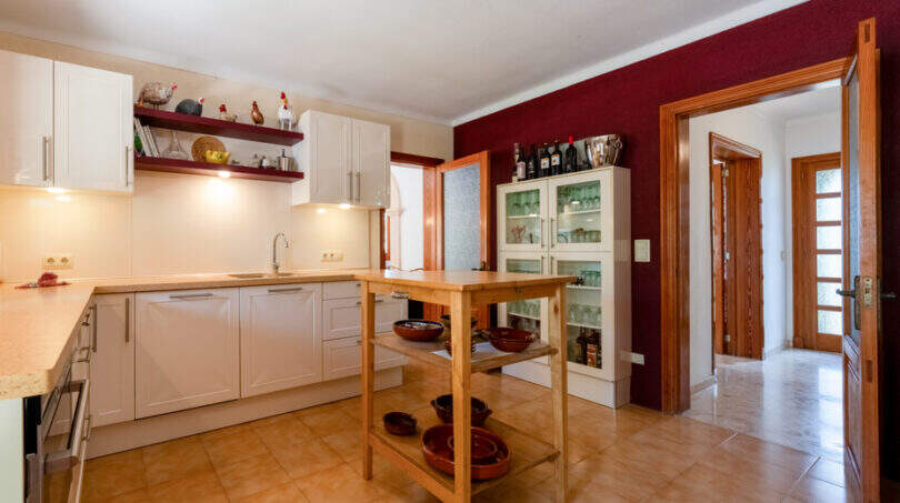 Amplia cocina de la villa de Cala Murada con isla de cocina, armarios blancos y una pared de acento en color burdeos