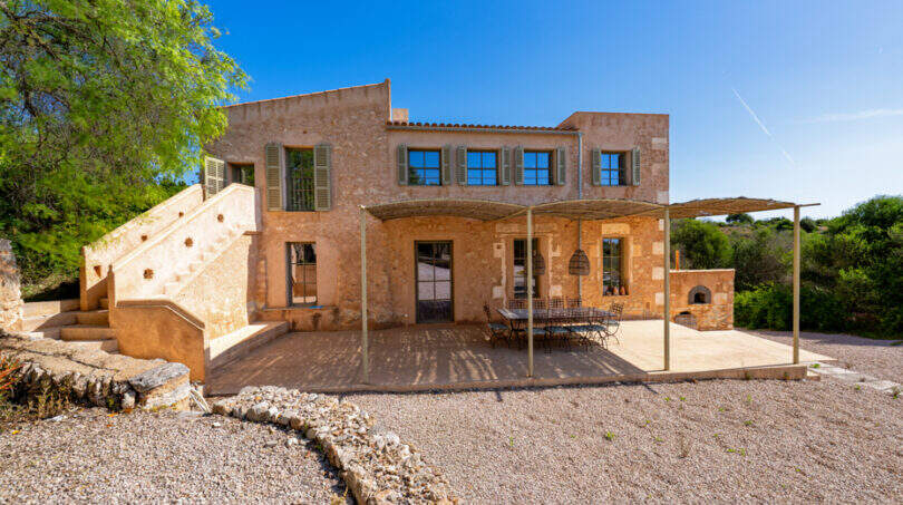 Rustikale Sandsteinfinca in Cales de Mallorca mit überdachter Terrasse, Grillplatz und Außentreppe unter blauem Himmel.