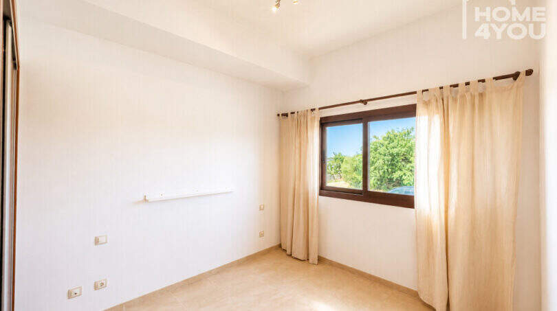 Helles leeres Schlafzimmer mit Fliesenboden, Einbauschrank und Fenster mit Leinenvorhängen, Blick ins Grüne