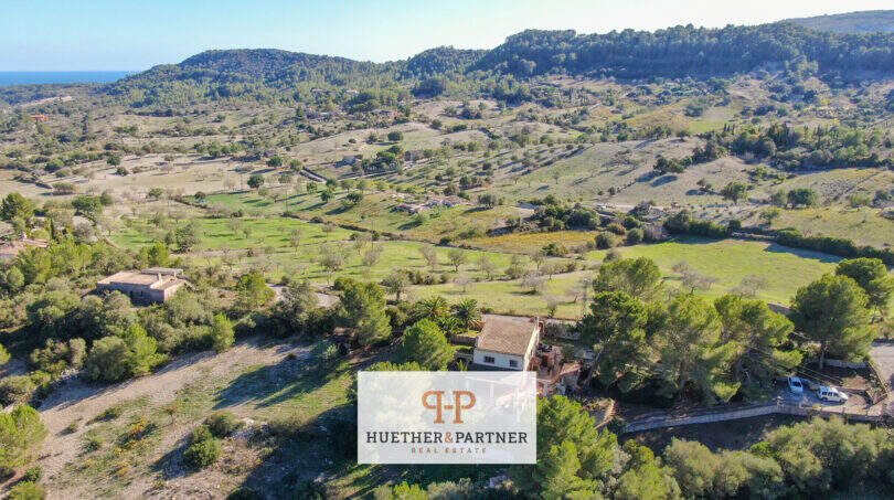 Luftaufnahme einer Finca in Son Macià, umgeben von grünen Hügeln, Bäumen und Feldern mit Meerblick.