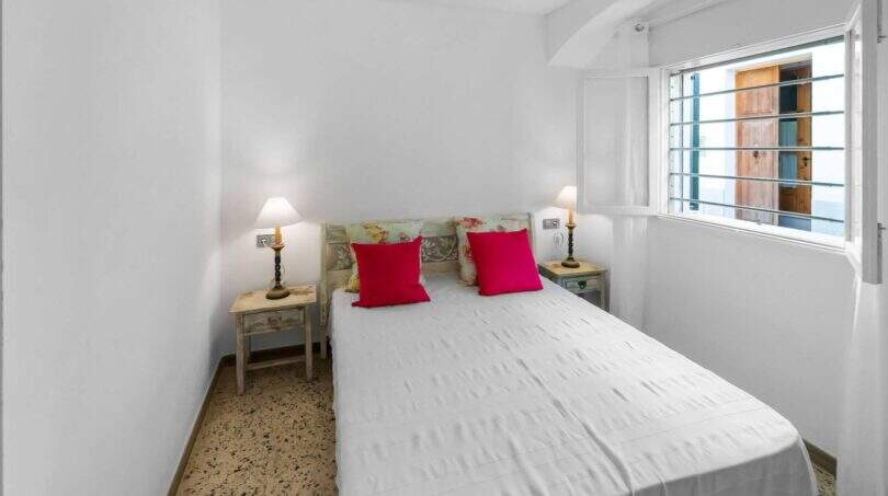 Luminoso dormitorio con cama doble, cojines rojos, cabecero de madera y ventana con vista al exterior en la casa de ciudad Felanitx