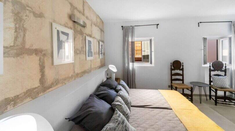 Dormitorio con pared de piedra natural, cama doble, colcha amarilla y sillas antiguas de madera en la casa de ciudad Felanitx