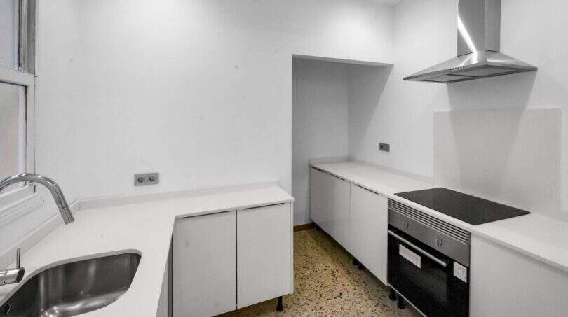 Moderna cocina blanca con horno empotrado, placa de inducción, campana extractora de acero inoxidable y suelo de terrazo en la casa de ciudad Felanitx