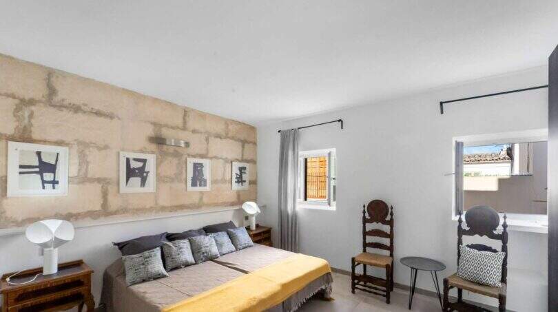 Dormitorio con pared de piedra natural, cama doble con colcha amarilla y sillas de madera antiguas en la casa urbana Felanitx