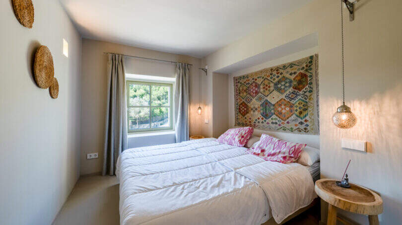 Helles Schlafzimmer der Finca in Esporles mit Doppelbett, buntem Wandteppich und rustikalem Holzbeistelltisch.