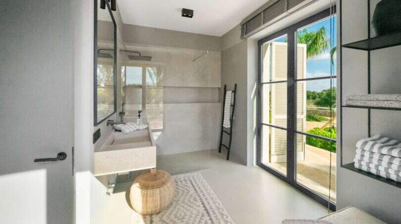 Modernes Badezimmer der Finca bei Portocolom mit Walk-in-Dusche, Naturstein-Waschtisch und Ausblick ins Grüne.