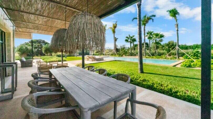 Überdachte Außenterrasse der Finca Portocolom mit Holztisch, Rattanstühlen, Palmen und Pool im Hintergrund
