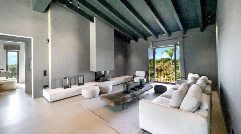 Helles Wohnzimmer der Finca Portocolom mit weißem Sofa, Kamin, Holzbalkendecke und Ausblick auf Palmen
